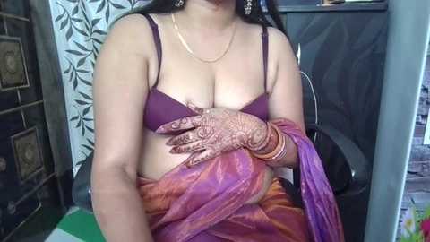Video of a South Asian woman with medium brown skin, wearing a purple bra, hennaed hands, and a colorful sari. She sits in a room with patterned curtains and a wooden cabinet.