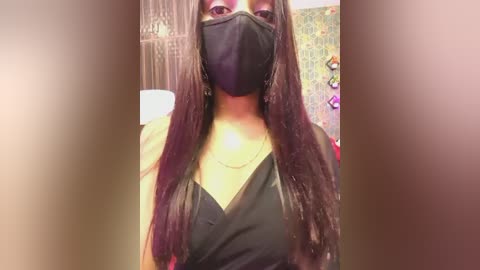 Video of a Black woman with long, straight hair wearing a black mask, black top, and necklace, indoors with a blurred background featuring a chandelier and colorful decor.