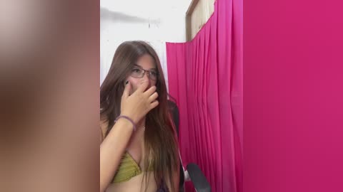 Video of a young woman with long brown hair, wearing glasses and a green bra, hiding her mouth with her hand, standing in a pink-walled changing room with a mirror.