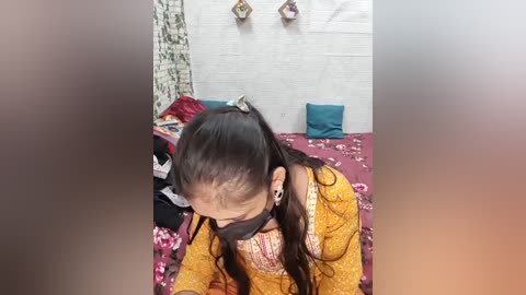 A video of a young South Asian woman with long black hair in a ponytail, wearing a yellow floral kurta, black face mask, and black earplugs, kneeling on a bed with a pink floral duvet.