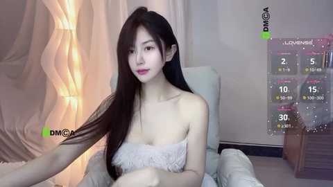Video of an East Asian woman with long black hair, pale skin, and small breasts, sitting on a white bed, wearing a white lace bralette. Background includes a curtain and a virtual reality headset display.