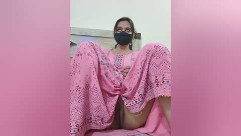 Media: A video of a South Asian woman with medium skin tone, wearing a pink lace dress, black mask, and sitting with legs spread, revealing her genitals.