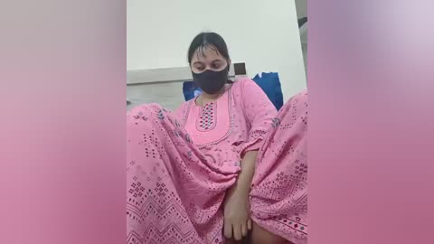 A video of a South Asian woman with medium skin tone, wearing a pink, patterned dress, black mask, and sitting with legs spread, in a dimly lit room.