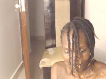 Video of a topless Black woman with short dreadlocks, standing in a narrow hallway with beige walls, a wooden door, and a beige chair.