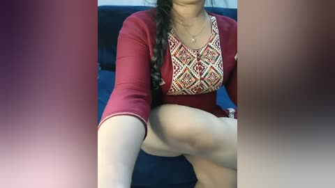 Video of a woman with light skin, long black hair in a braid, wearing a maroon top with intricate white and red embroidery, sitting on a dark blue sofa, legs visible.