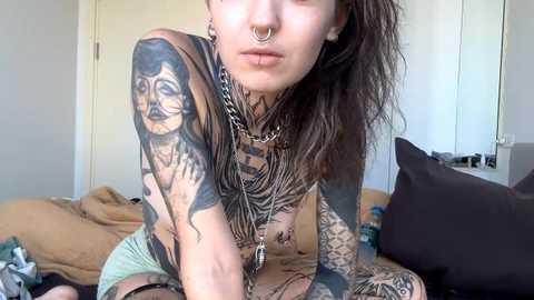 Video of a tattooed, pale-skinned person with long brown hair, wearing a chain necklace, nose ring, and a septum ring, sitting on a bed with brown sheets and pillows, in a plain, beige room.