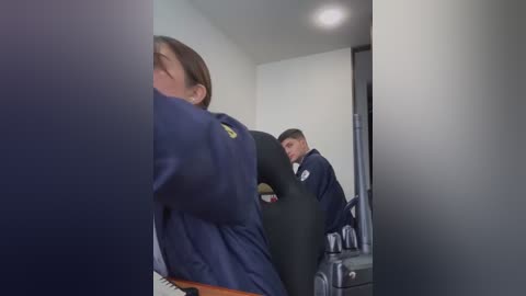 A video shows a woman in a navy-blue jacket with a yellow emblem, her face obscured by a blurred door frame, and a man in a dark suit standing beside her in a dimly lit hallway.