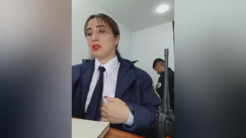 Video of a woman in a navy blue suit and red lipstick, holding a pen, in a modern office with a man in uniform standing in the background.