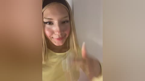 Video of a smiling young woman with straight blonde hair, light skin, and a yellow top, partially obscured by a white wall and a blurry arm.