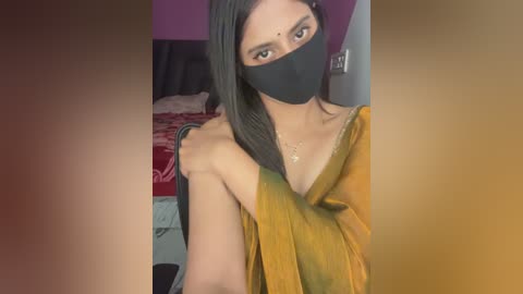Video of a young South Asian woman with a fair complexion, wearing a black mask, mustard-yellow sari, and a delicate necklace. She is indoors, with a bed in the background.