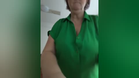 Video of a middle-aged woman with short, dark hair wearing a green button-down shirt. She stands indoors with a blurred, green-tinted background.