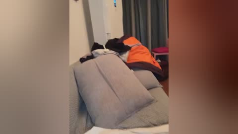 Video of a messy bedroom with a person in a bright orange and black outfit sleeping on a bed with grey and white pillows and sheets. Dark curtains cover a window in the background.