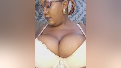Video of a dark-skinned woman with medium-sized breasts in a white lace bra, wearing glasses and a headscarf, with floral wallpaper in the background.