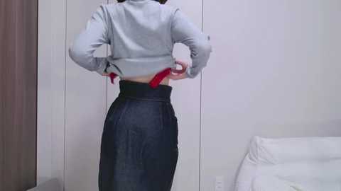 Video of a woman in a light grey sweatshirt and high-waisted dark blue skirt, adjusting her red belt in a minimalist, modern bedroom with white walls and a bed.