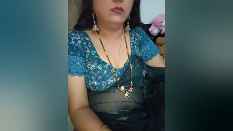 Media: Video of a woman with medium brown skin and long black hair, wearing a blue floral top with lace sleeves and a black sheer dress underneath, adorned with gold necklaces and earrings, holding a pink plush toy.