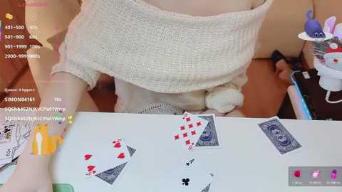 Video of a woman wearing a white off-shoulder sweater, playing cards, and a plush toy on a table.
