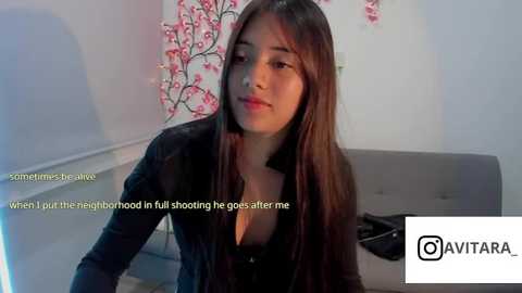 Video of a young Asian woman with long dark hair, wearing a black top, seated on a gray couch in a room with pink cherry blossom wall art. Text overlays her face.