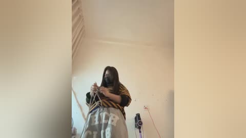 A video of a young person with long brown hair, wearing a yellow and black striped shirt and gray pants, using a rope to climb a white wall in a minimalist room with beige walls and a red electrical cord.