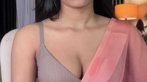 Video of a woman with medium skin tone, wearing a gray ribbed tank top with a pink shawl draped over her left shoulder, in a softly lit room with orange lamp and dark curtains.