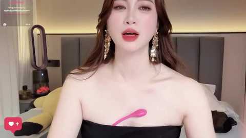 Video of a young Asian woman with long brown hair, wearing a strapless black dress, and large gold earrings, lying on a bed with a grey headboard, a pink vibrator in her hand, in a modern bedroom.