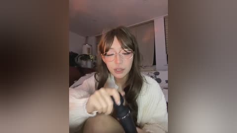 A young woman with long brown hair and glasses, wearing a white sweater, sits on a bed, holding a black object. The room has white walls, a dark blanket, and a lamp.