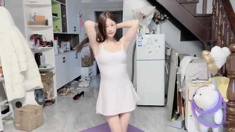 Video of a young Asian woman with long brown hair, wearing a white dress, arms raised, standing in a cluttered, messy living room with white cabinets, boxes, shoes, and toys scattered around.