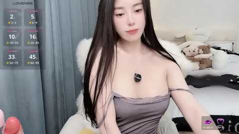 Video of an Asian woman with long black hair, wearing a strapless lavender top, sitting in a bedroom with a white bed and stuffed animals, displaying a virtual dating app interface.
