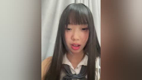 A video of a young Asian girl with long black hair, wearing a white collared shirt and gray vest, standing against a white curtain background.