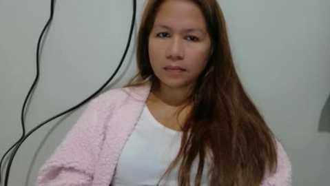 A video of a young Asian woman with long brown hair, wearing a white top and light pink fuzzy robe, standing against a plain white wall with a black electrical cord hanging nearby.