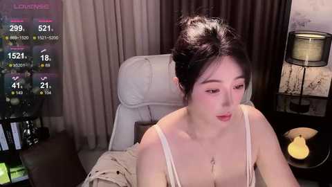 Video of an Asian woman with pale skin, dark hair in a messy bun, wearing a white tank top, sitting in a white chair, indoors with a digital clock display, and a lamp in the background.