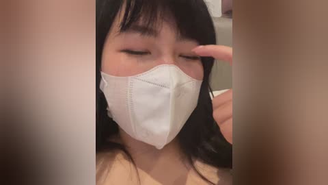 Video of an East Asian woman with long black hair, wearing a white face mask, eyes closed, and touching her face with her right hand. Background is blurry, suggesting indoor setting.
