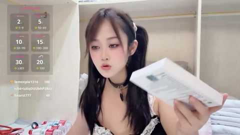 A video of an East Asian woman with long black pigtails, wearing a black dress with lace trim, holding a magazine. Background shows a white closet and a calendar.