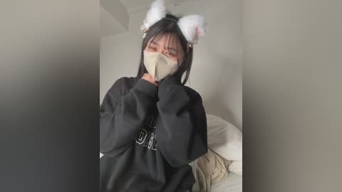 A video of an Asian woman in a black hoodie, white cat ears, and beige face mask, standing in a minimalist, dimly lit room with white walls and a bed.