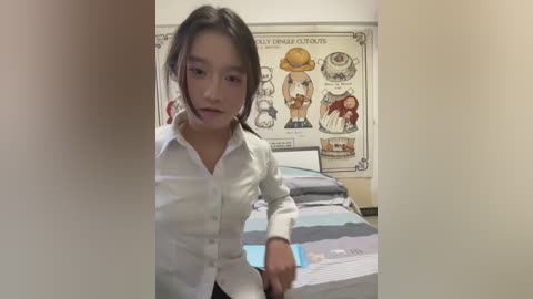 Video of a young Asian woman with medium-length dark hair, wearing a white button-up shirt, standing in a bedroom with a bed covered in a blue and gray striped blanket. The room features a wall poster with cartoon characters and a white wooden headboard.