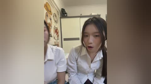 A candid video of a young Asian woman with long black hair, wearing a white blouse and looking into a mirror, in a cluttered room with posters and a closet.