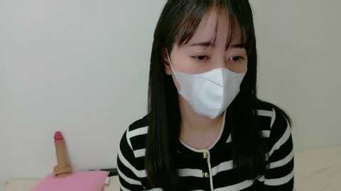 Video of an Asian woman with long black hair, wearing a white surgical mask, black and white striped shirt, and a beige bedspread. Background includes a pink dildo and a pink book.