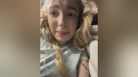 Video of a young blonde woman with light skin, wearing a light blue shirt, lying on a bed. Her hair is styled in loose braids. The background is blurred.