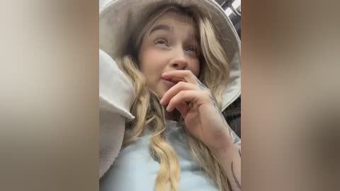 Video of a blonde woman with wavy hair, wearing a beige hat and light blue shirt, with her fingers to her lips, in a blurred, beige-toned background.