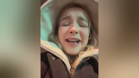 A candid video captures a young blonde girl with closed eyes and a slightly open mouth, wearing a beige hooded jacket, against a blurred, beige background.