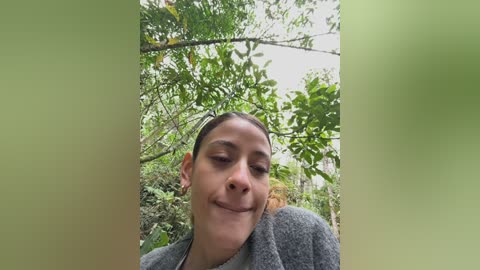 Video of a young woman with olive skin, brown eyes, and dark hair pulled back, smiling in a lush, green forest setting with trees and foliage in the background.