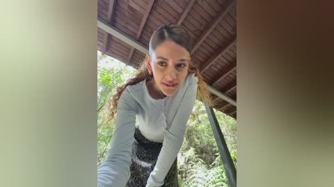 Video of a young woman with light brown skin, long wavy hair in braids, wearing a light blue long-sleeve top and black patterned skirt, leaning forward under a wooden-roofed structure with lush greenery in the background.