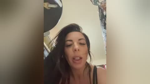 A video of a woman with long black hair, wearing a black top, seen through a doorway, in a dimly lit room with hanging clothes.