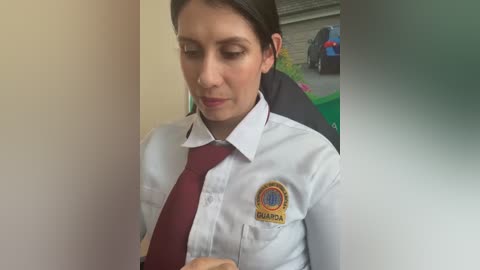Video of a young woman with brown hair, wearing a white school uniform shirt with a maroon tie and a badge, standing in a hallway next to a black trash can and a green bin.
