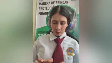 Video of a woman with long black hair, wearing a white shirt and maroon tie, standing in front of a Spanish security guard sign, focusing intently on her phone.
