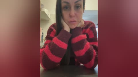 Video of a woman with long brown hair, wearing a red and black striped sweater, leaning forward, looking serious. Background shows a blurred, neutral-toned room.
