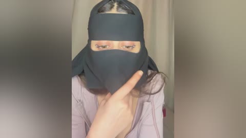 Video of a person wearing a black face mask, with their hand under the mask, revealing their face. They have light skin and brown hair. Background is a beige curtain.