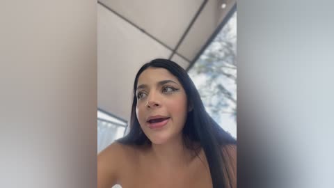 Video of a smiling, fair-skinned woman with long, straight black hair, wearing a strapless top, standing indoors with a blurred background.