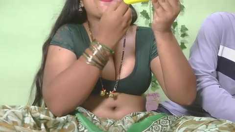 Video of a dark-skinned woman with long black hair, wearing a low-cut green top and green patterned skirt, eating a yellow popsicle, in a green room with a patterned wall.