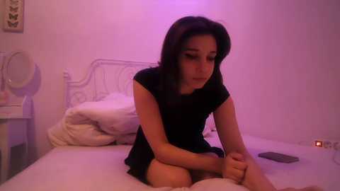 Video of a young woman with long dark hair, sitting on a bed in a dimly lit, softly purple-lit room, wearing a short black dress.