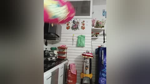 Video of a small, cluttered kitchen with white walls, a microwave, stove, and a pink-and-white spiral-shaped object in mid-air. Background includes colorful floral decorations, green bags, and blue clothing hanging.